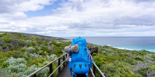 Hiker wearing large backpack full of gear walking along wooden footpath on the Cape to Cape track in Western Australia
