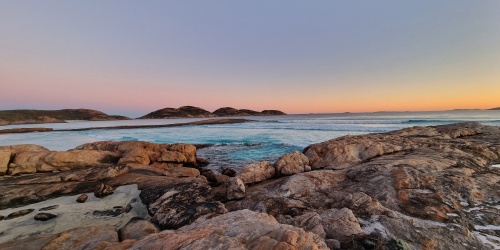 Rocky shoreline at Lucky Bay in Cape Le Grand National Park at sunset, with rounded granite rocks in the foreground, clear turquoise water and gentle waves, and low islands on the horizon beneath a soft pink and orange sky