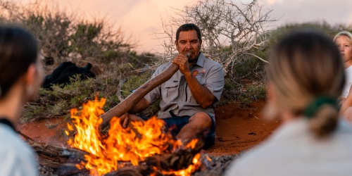 A man plays the didgeridoo in front of a glowing campfire