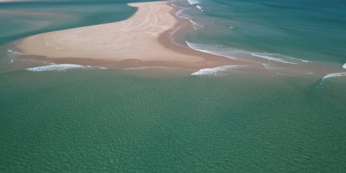 Aerial view of a pristine coastal sandbar extending into clear turquoise waters, with gentle waves breaking along the shoreline. White sandy beaches and shallow, aquamarine channels surround the sand spit, while low dunes stretch across the distant coastline beneath a blue sky dotted with scattered clouds.