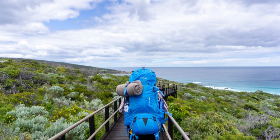 Hiker wearing large backpack full of gear walking along wooden footpath on the Cape to Cape track in Western Australia