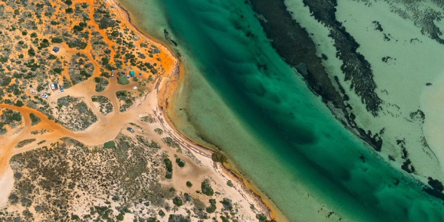 Aerial view of a rugged coastline with orange sand dunes meeting clear turquoise water and a narrow tidal channel.