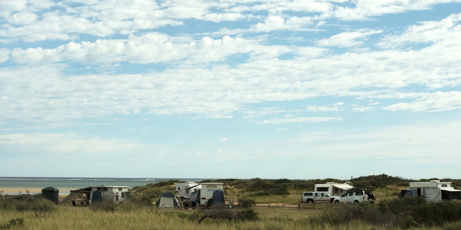 Several vehicles and caravans are in a beachside campground. Light clouds are in the blue sky above.