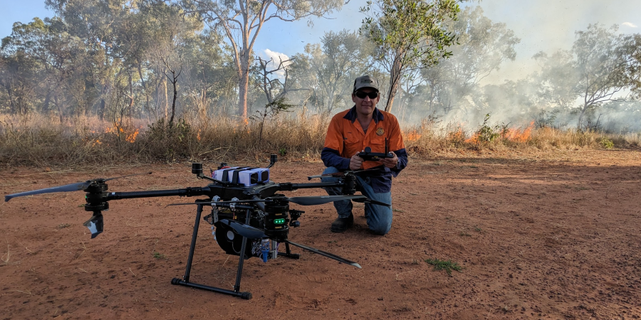 A man kneeling behind a large drone with trees and bush in the background.