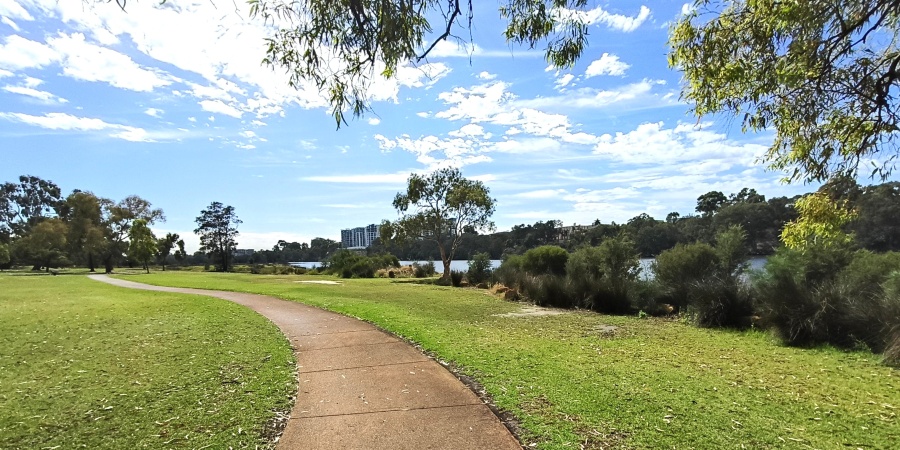 Curving paved walking path through green parkland beside a river, with trees and shrubs along the foreshore, distant buildings across the water, and a bright blue sky with scattered clouds overhead.