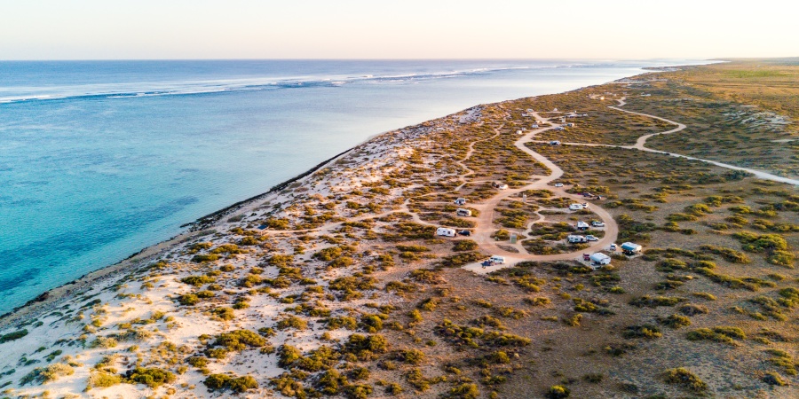Aerial view of a Kurrajong Campground in Cape Range National Park with sandy tracks winding through low scrubland, dotted with parked caravans and vehicles beside a calm blue ocean and a long sandy shoreline.
