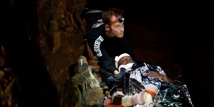 Rescuer assisting an injured person wrapped in a foil blanket in a dark, narrow gorge at night.