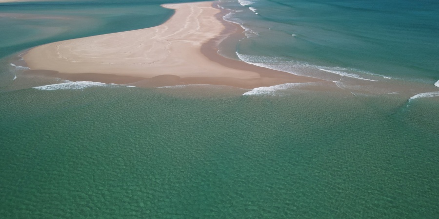 Aerial view of a pristine coastal sandbar extending into clear turquoise waters, with gentle waves breaking along the shoreline. White sandy beaches and shallow, aquamarine channels surround the sand spit, while low dunes stretch across the distant coastline beneath a blue sky dotted with scattered clouds.