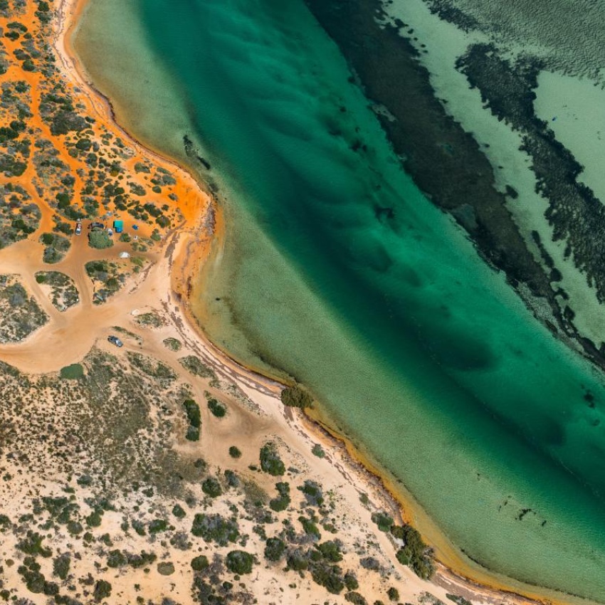 Aerial view of a rugged coastline with orange sand dunes meeting clear turquoise water and a narrow tidal channel.