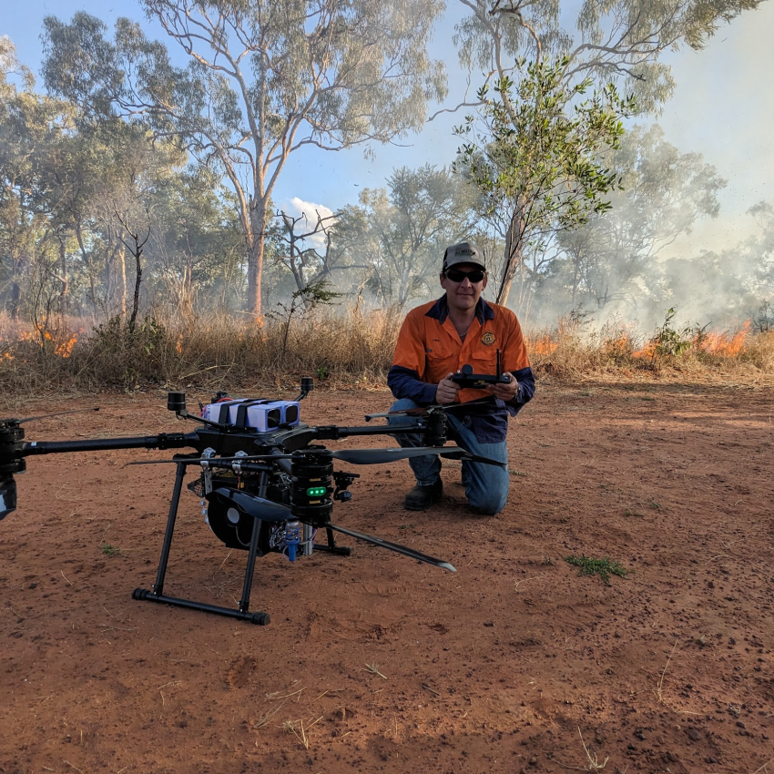 A man kneeling behind a large drone with trees and bush in the background.
