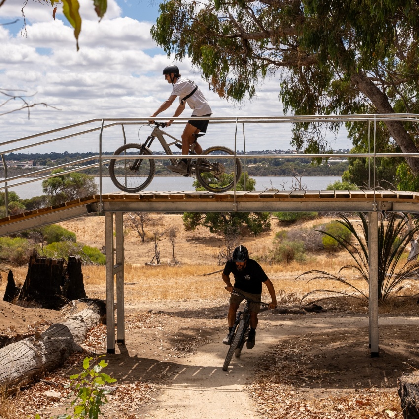 Two mountain bikers ride on a sunny trail; one crosses a bridge, and the other navigates the path below. 