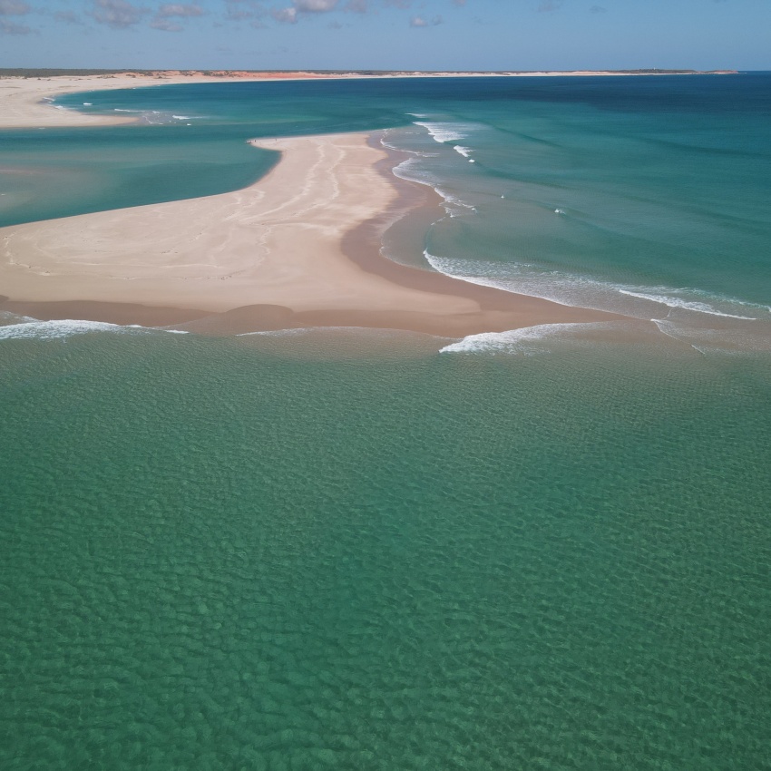 Aerial view of a pristine coastal sandbar extending into clear turquoise waters, with gentle waves breaking along the shoreline. White sandy beaches and shallow, aquamarine channels surround the sand spit, while low dunes stretch across the distant coastline beneath a blue sky dotted with scattered clouds.