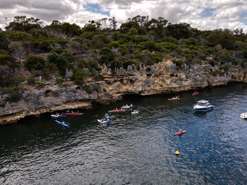 Aerial view of kayakers paddling along a dark river beside a rugged limestone cliff topped with dense native bushland, with a small motorboat nearby under a cloudy sky.