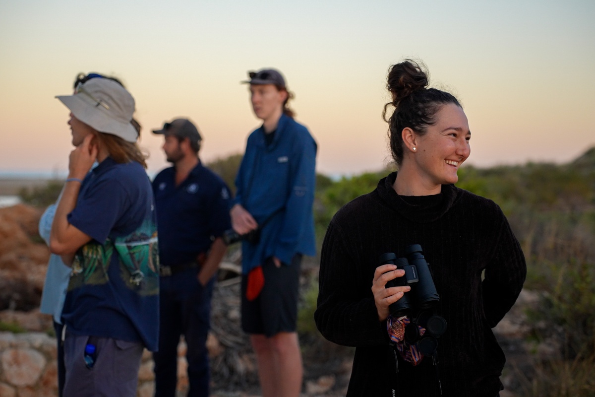 A woman holding binoculars smiles as the sun sets outside. There are other figures blurred in the background. 