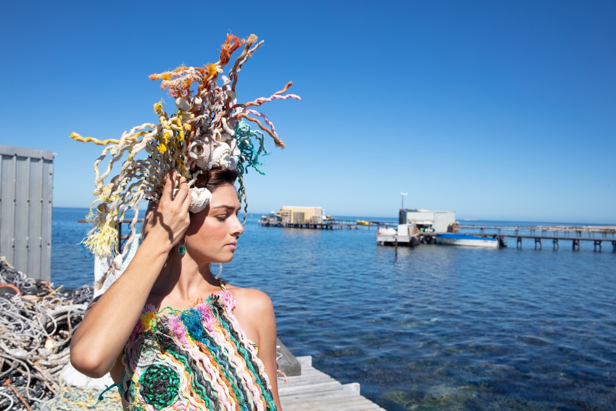 A woman wears art made from marine debris, including a halter top and a headpiece. She stands on a jetty in front of the ocean. 