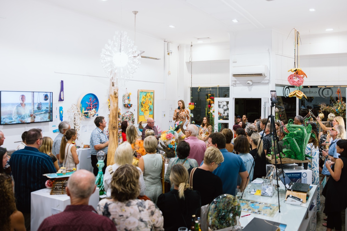 Community gathers at the Flotsam and Jetsam art event. The crowd faces Pia who is presenting on stage, surrounded by art.