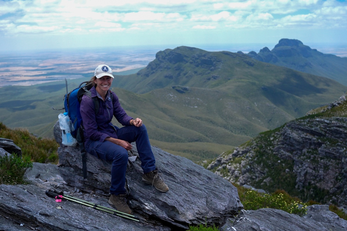 A smiling woman sits on a mountain peak with more mountains in the background