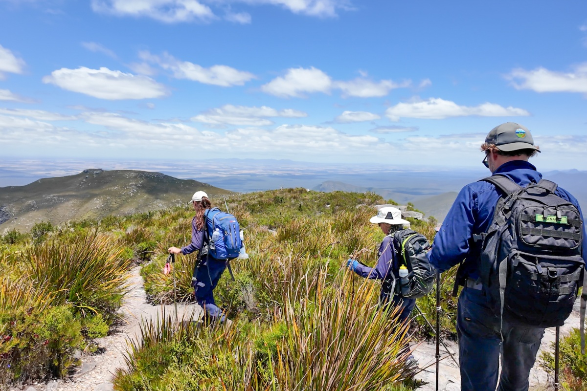 Three people hike through the mountaintop with hiking gear in the sunshine