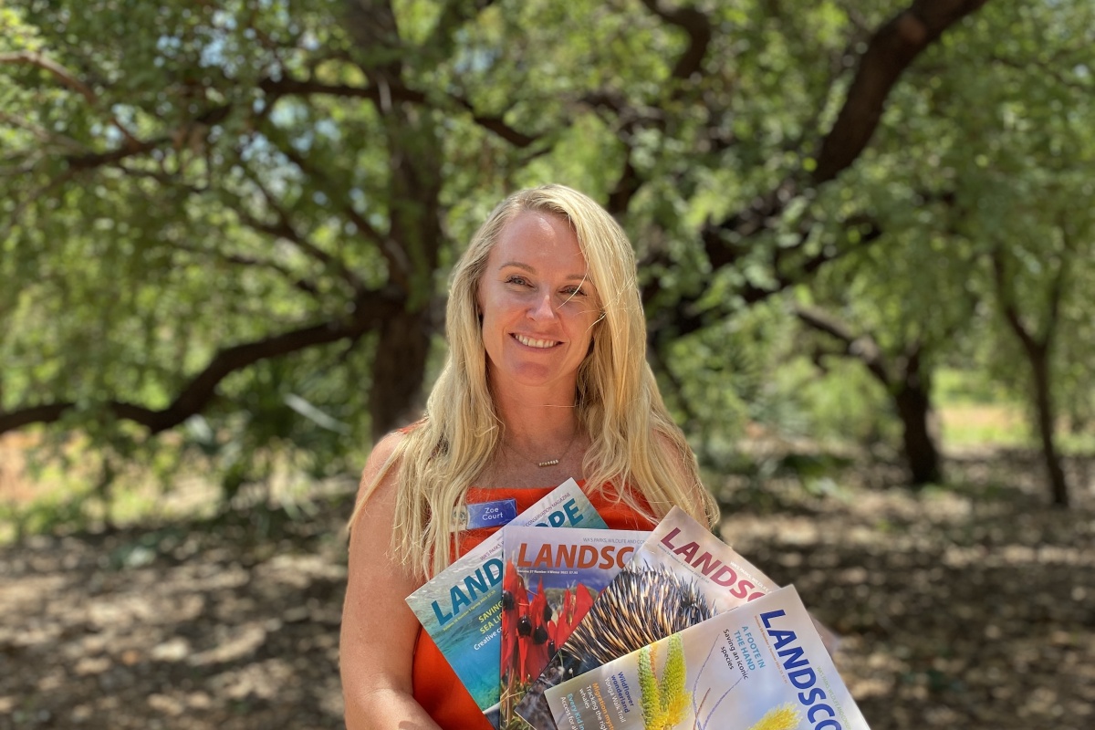 A blonde female teacher stands with issues of a magazine outside in front of a tree