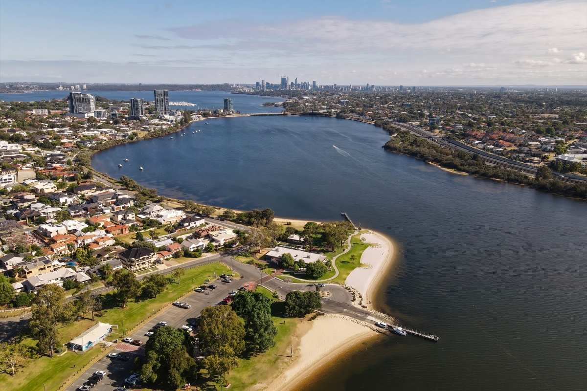 Aerial view of the Swan River curving around a suburban foreshore with sandy beaches, parkland, and residential areas, with Perth’s city skyline visible in the distance and boats scattered across the water.