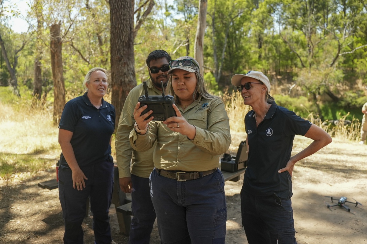 Four people standing in a bushland setting gathered around a handheld controller, likely for a drone, with one person operating the device while the others observe; a drone sits on the ground nearby among dry grass, trees, and dappled sunlight