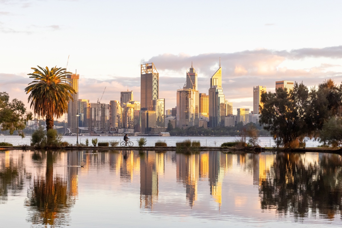 Perth city skyline at golden hour viewed across calm water, with modern high-rise buildings lit by warm sunlight, reflections in the foreground, a cyclist riding along a path by the river, and a large palm tree on the left framing the scene.