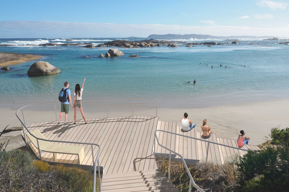 Elevated wooden viewing platform with stairs and railings overlooking a calm, turquoise ocean dotted with large rounded rocks. Several people are in the water swimming and wading near the shore, while a few people sit or stand on the steps and deck, one pointing out toward the sea. Waves break gently farther out, and low hills or coastline stretch across the horizon under a clear blue sky.