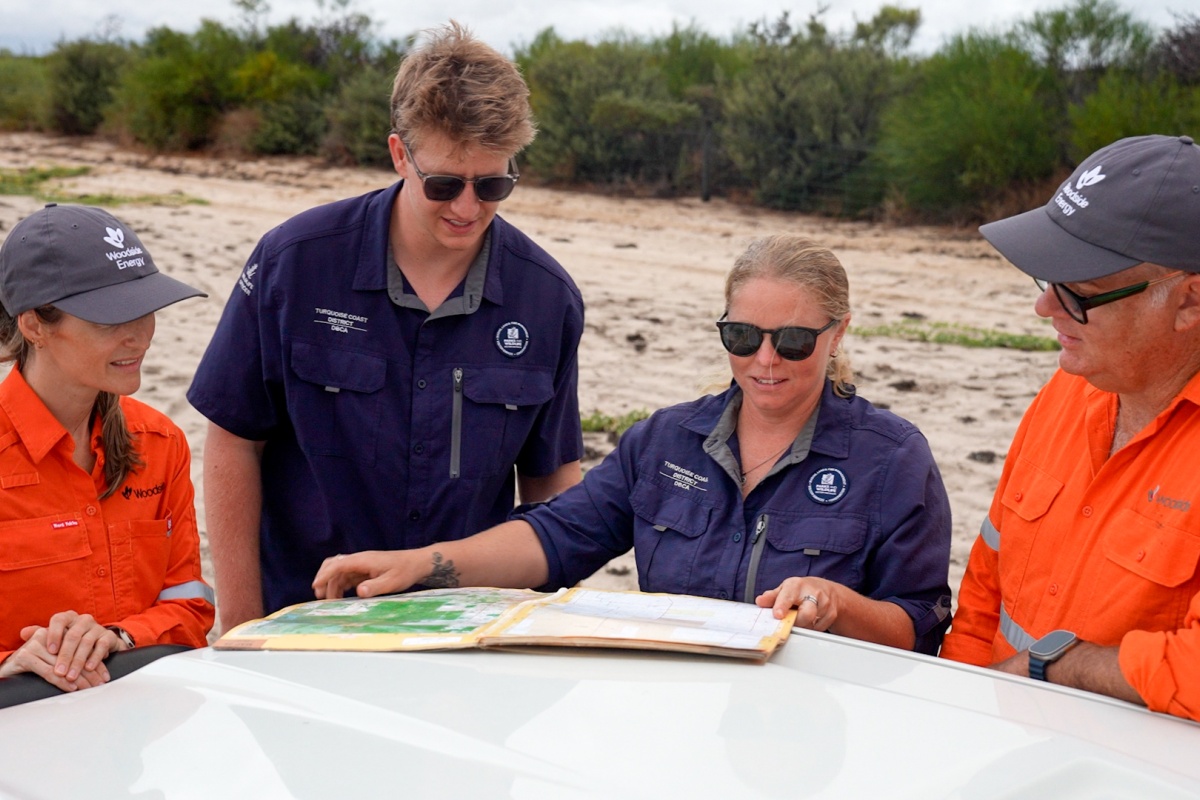 Four field staff in high-visibility orange and navy uniforms stand around a vehicle in a sandy bushland area, reviewing a large map spread across the vehicle’s surface. Low scrub vegetation surrounds the group under a cloudy sky, with Woodside Energy and Parks and Wildlife logos visible on clothing.