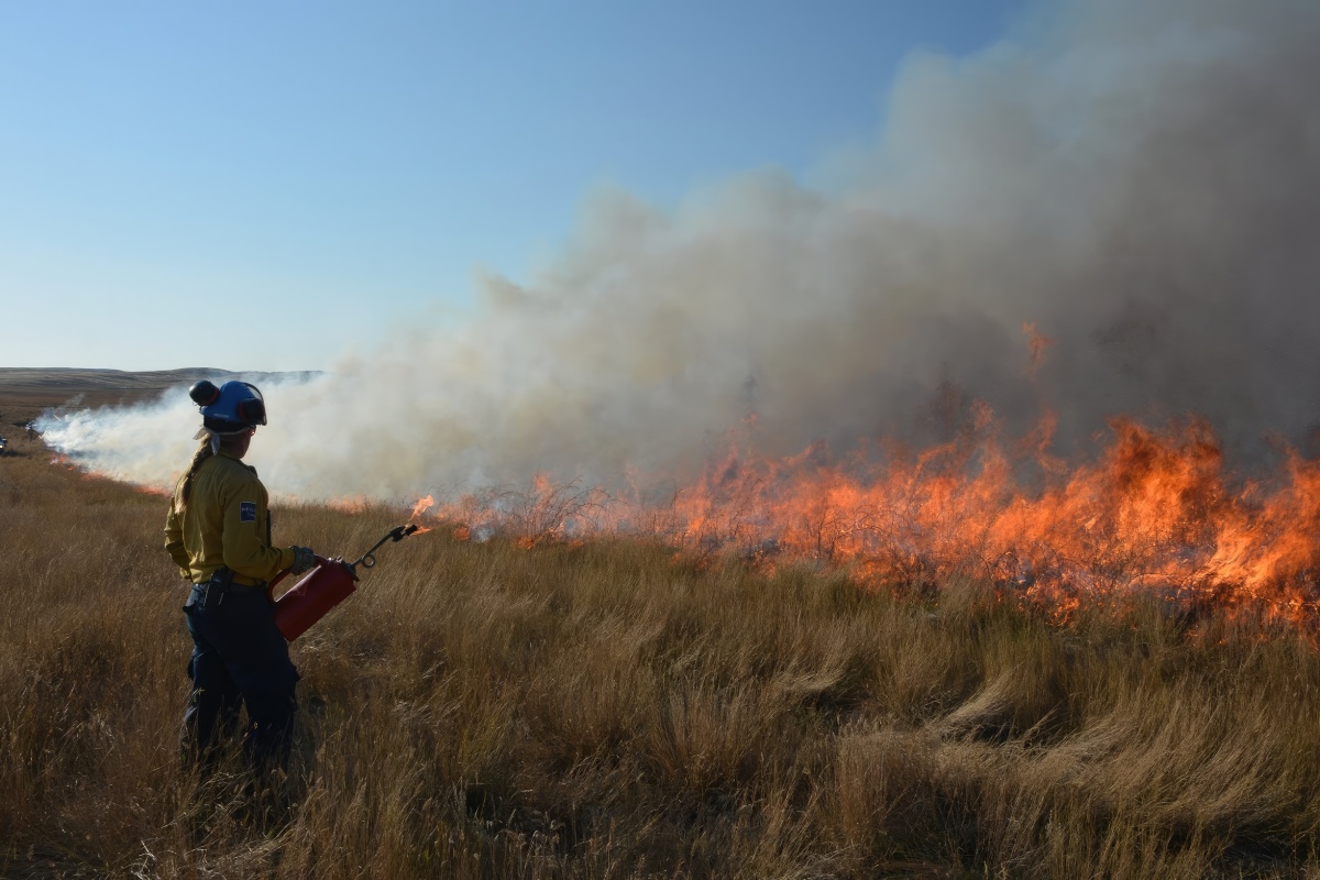 A woman stands in firefighting gear in front of a burning field doing prescribed burns