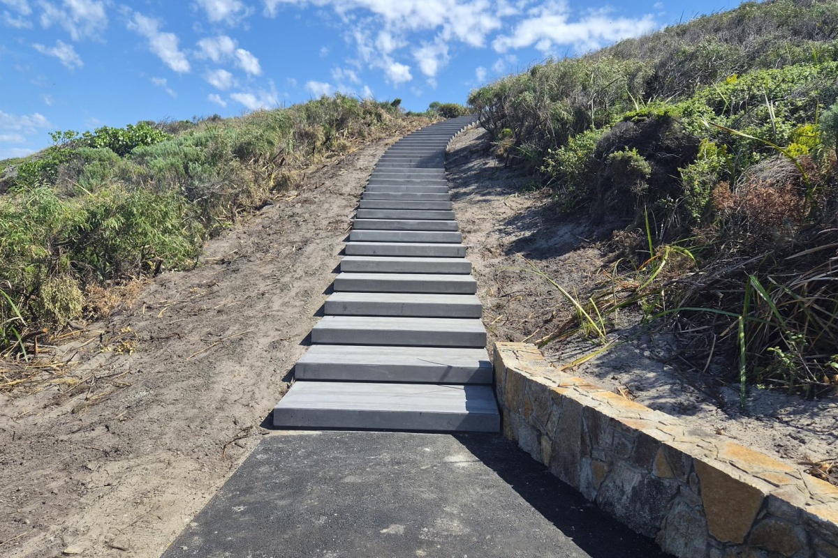 A wide timber staircase leads up a sandy coastal hillside covered in native shrubs and grasses. A low stone retaining wall lines one side of the path, with blue sky and scattered white clouds overhead